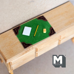 May include: A miniature wooden desk with three drawers, a green felt writing surface, a white piece of paper, a pencil, and a small yellow box. The desk is decorated with brown corner accents.