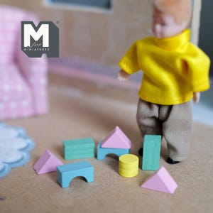 May include: A miniature dollhouse scene featuring a doll in a yellow shirt and tan pants. Colorful wooden blocks in shades of pink, blue, green, and yellow are scattered on the floor. The logo "M for miniatures" is visible in the upper left corner.