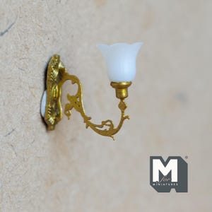 May include: A miniature wall sconce with a gold-colored, ornate arm and a white, tulip-shaped shade. The sconce is mounted on a white base and is designed for a dollhouse or miniature setting. The overall design is vintage-inspired.