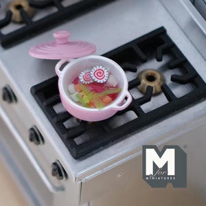 May include: A miniature pink pot with a lid, filled with colorful vegetables and two pink and white swirl decorations, sits on a black stovetop.