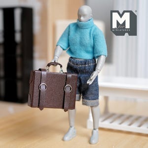May include: Miniature brown briefcase with silver-colored hardware. The briefcase has a handle and two straps with buckles. The briefcase is held by a miniature figure wearing a blue sweater and denim shorts. The logo "M for MINIATURES" is in the upper right corner.