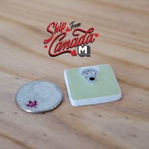 May include: A miniature green and white bathroom scale with a silver dial. The scale is next to a Canadian coin with a red maple leaf.