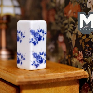 Op de afbeelding: Een witte keramische vaas met een blauw-wit bloemenpatroon. De vaas is rechthoekig en heeft een vierkante basis. Hij staat op een houten oppervlak.