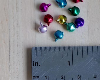 May include: A collection of ten small, metallic jingle bells in various colors including red, blue, green, purple, yellow, and pink, positioned above a metal ruler showing measurements in inches and centimeters for scale.