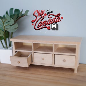 May include: A miniature wooden cabinet with three drawers and open compartments. One drawer is partially open, revealing the interior. The cabinet is light-colored and sits on a wooden surface. A potted plant is in the background.