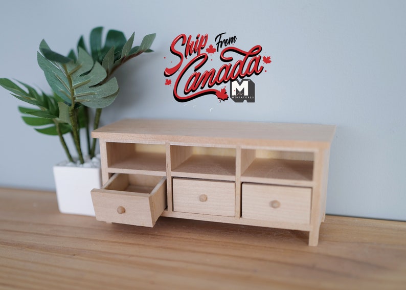 May include: A miniature wooden cabinet with three drawers and open compartments. One drawer is partially open, revealing the interior. The cabinet is light-colored and sits on a wooden surface. A potted plant is in the background.