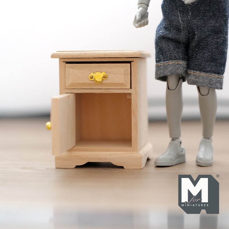 Peut inclure: Une petite table de chevet en bois avec un tiroir et une porte. La porte est ouverte et le tiroir est ferm&eacute;. La table de chevet est en bois clair et a une poign&eacute;e dor&eacute;e.