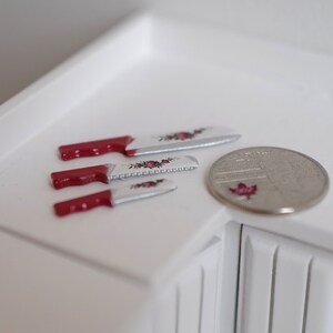 May include: Three miniature knives with red handles and floral designs on the blades, next to a Canadian quarter.