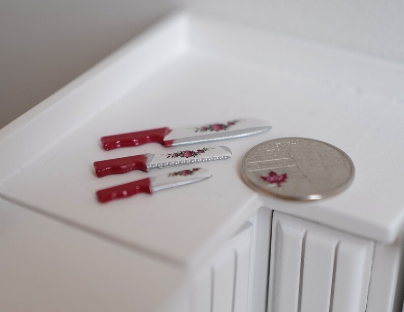 May include: Three miniature knives with red handles and floral designs on the blades, next to a Canadian quarter.