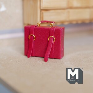 May include: A miniature red suitcase with gold-colored hardware, including a handle and ring details. The small luggage has two straps with gold rings and is designed for dollhouses or miniature displays. The suitcase is a vibrant red color.
