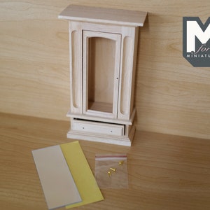 May include: A wooden dollhouse cabinet with a glass door and a drawer. The cabinet is unfinished and comes with two sheets of paper and a bag of small gold-colored tacks.