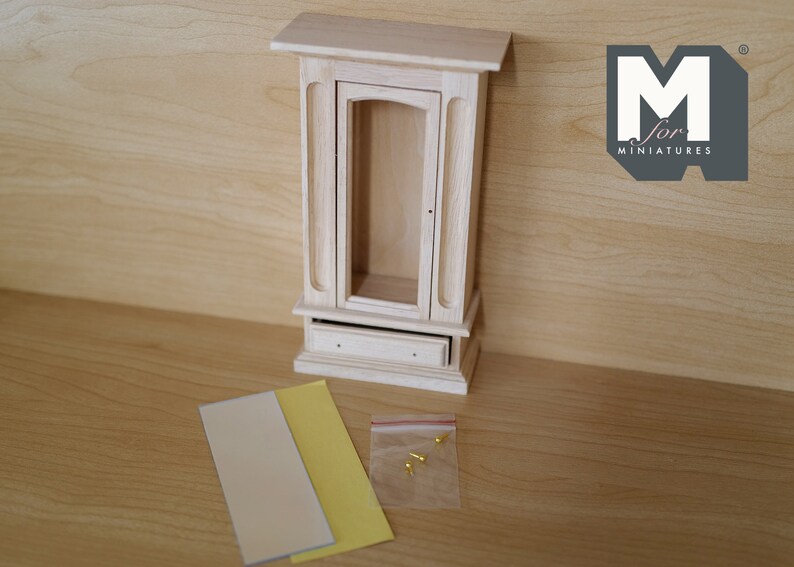 May include: A wooden dollhouse cabinet with a glass door and a drawer. The cabinet is unfinished and comes with two sheets of paper and a bag of small gold-colored tacks.