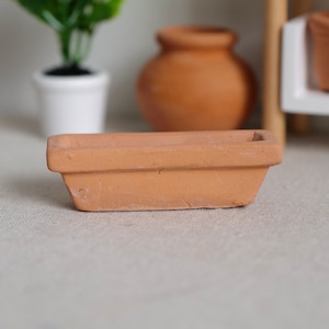 Op de afbeelding: Een kleine, rechthoekige terracotta plantenbak met een licht taps toelopend ontwerp. De ongeglazuurde kleipot heeft een natuurlijke, aardse tint en een rechthoekige opening. Andere terracotta potten en een witte pot met een plant staan op de achtergrond.