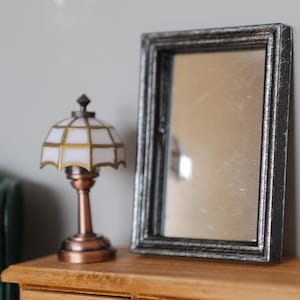 Könnte beinhalten: Miniatur-Lampe mit weiß-gold gemustertem Schirm und bronzefarbenem Sockel. Daneben ein kleiner rechteckiger Spiegel mit silberfarbenem Rahmen. Die Objekte stehen auf einer Holzoberfläche, möglicherweise für ein Puppenhaus.