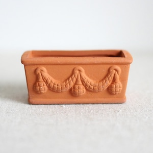 Può includere: Piccolo vaso rettangolare in terracotta. Il vaso presenta un motivo decorativo a ghirlanda con tre forme pendenti. Il vaso è di un caldo colore arancione terroso ed è adatto a piccole piante.