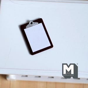 May include: Miniature brown clipboard with a silver clip holding a blank white paper. The clipboard is rectangular and has a dark brown border. The image is of a miniature item, likely for a dollhouse or craft project.