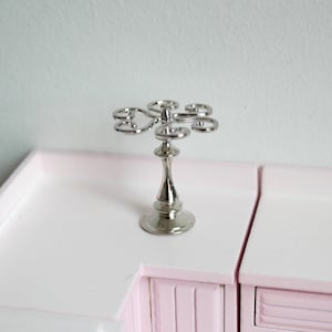 May include: Silver metal decorative stand with a round top and five curved arms. The stand is on a pink surface.