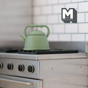 May include: A green miniature tea kettle with a handle and spout sits on a black stovetop burner. The stovetop is silver and has four knobs. The background is a white tiled wall with the text "M for Miniatures" in black.
