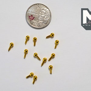 May include: A collection of ten gold-coloured metal knobs with a round head and a small pin on the bottom. The knobs are scattered on a white surface.