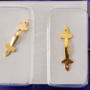 May include: Two gold-colored metal picture frame hangers in clear plastic packaging. One hanger is straight, the other is angled. Each has a decorative top and bottom with a hole for hanging.