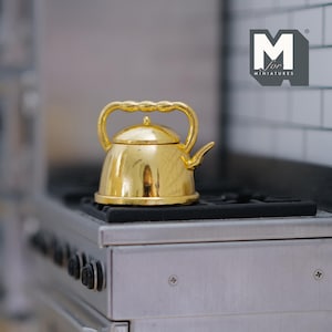 May include: A miniature gold-coloured kettle with a curved handle and spout, sitting on a black stovetop. The kettle has a shiny, reflective surface. The image includes the text "M for MINIATURES".