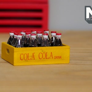 May include: A yellow wooden crate with the words "Cola Cola Drink" printed on the front. The crate is filled with miniature glass bottles of cola.