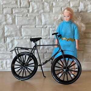 May include: A black bicycle with a white doll standing next to it. The doll is wearing a blue dress with gold trim.
