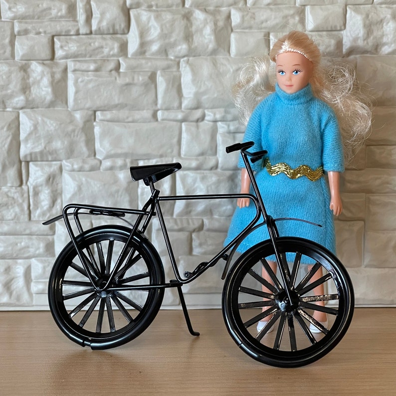 May include: A black bicycle with a white doll standing next to it. The doll is wearing a blue dress with gold trim.