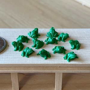 Pode incluir: Uma coleção de doze figuras de sapo de plástico verde. Os sapos estão todos em poses diferentes, alguns estão sentados, outros em pé e outros pulando. Os sapos medem aproximadamente 2,5 cm de altura.