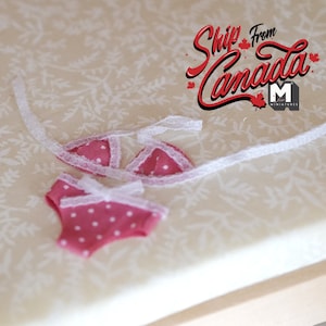 May include: A pink and white polka dot bikini with white lace trim. The bikini is made for a doll and is laying on a white fabric with a floral pattern.
