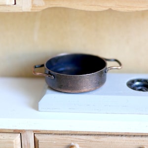 May include: A small, copper-colored pot with two handles sits on a white countertop. The pot is empty and has a dark interior.