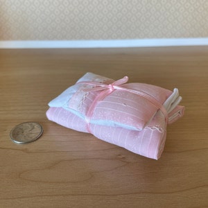 May include: A set of pink and white doll-sized bedding, tied with a pink ribbon. The bedding is made of a soft, patterned fabric and includes a pillow and a blanket.