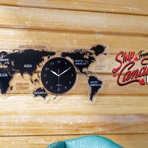 May include: A black and white world map clock with a black face and white hands. The clock is mounted on a wooden wall with the text "Ship From Canada" in red lettering.