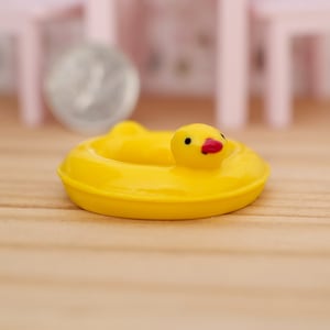 May include: A miniature yellow duck-shaped pool float with a red beak and black eyes. The toy is made of plastic and is designed for dollhouses or other miniature settings. The float is on a wooden surface.