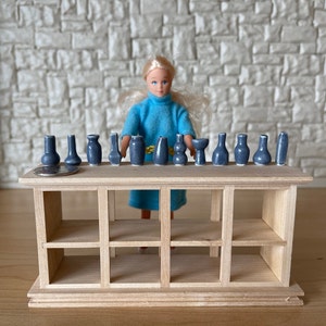 May include: A wooden shelf unit with 12 small blue ceramic vases arranged on the top shelf. A doll wearing a blue dress is standing in front of the shelf.