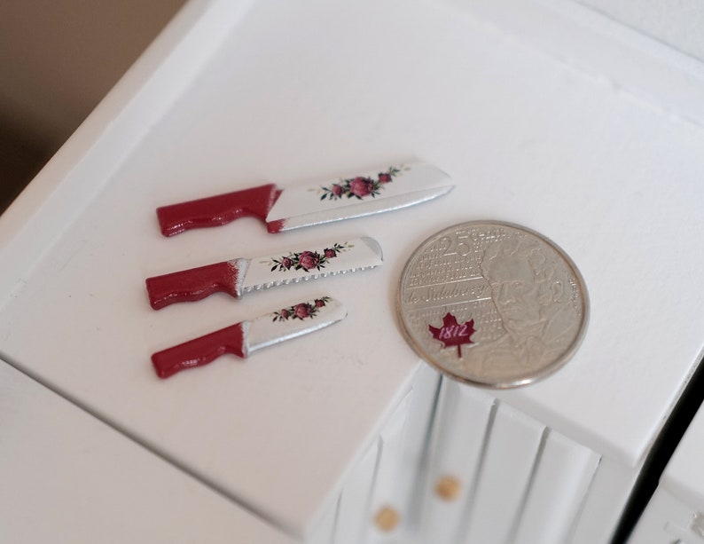May include: Three miniature knives with red handles and white blades decorated with pink roses. The blades are slightly serrated. A silver coin with a red maple leaf and the year 1812 is in the background.