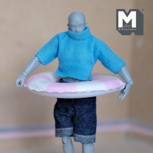 May include: A miniature figure wearing a light blue sweater, denim shorts, and a pink and white striped inflatable ring. The figure is a doll, and the image is of a miniature scene. The logo "M for MINIATURES" is in the upper right corner.