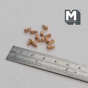 May include: A collection of small, light brown wooden miniature finials or decorative pegs, each featuring a rounded top and a tiered, cylindrical base. A metal ruler is positioned alongside the items, showing a scale in inches and centimeters.