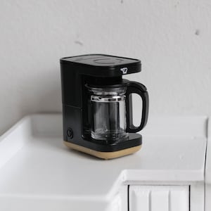 Pode incluir: Máquina de café preta com uma jarra de vidro e uma base bege. A máquina de café está em cima de uma bancada branca.