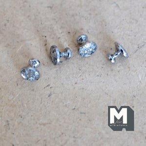 Peut inclure: Quatre petits clous en métal argenté avec des têtes ovales et des surfaces texturées. Chaque clou présente une courte tige cylindrique s'étendant à l'arrière de la tête. Le logo M for Miniatures est visible dans le coin inférieur droit.