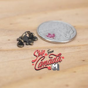 May include: A small, antique bronze-colored metal button featuring a fleur-de-lis design with a raised loop on the back, positioned next to a Canadian quarter coin for scale, which displays a red maple leaf graphic and the year 1812 on its surface.