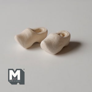Op de afbeelding: Twee kleine, ongeverfde houten klompen, ook wel Nederlandse schoenen genoemd, worden getoond op een wit oppervlak. De klompen zijn gemaakt van onbehandeld hout en hebben een afgeronde neus.