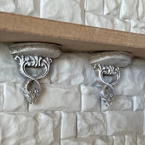 May include: Two ornate, distressed white shelf brackets with a decorative scroll design are mounted on a white brick wall. The brackets support a wooden shelf, creating a vintage or antique aesthetic.