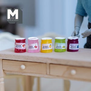 May include: Miniature jars of fruit preserves in red, pink, yellow, green, and purple. Each jar has a white lid and a label with the fruit type. The jars are arranged on a light wood surface. The jars are labeled "Strawberry jam", "Cherry jam", "Orange jam", "Kiwi jam", and "Blueberry jam".