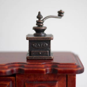May include: A small, antique-looking metal coffee grinder with a crank handle. The grinder is black with a copper-coloured trim and says "Cafe" on the front.