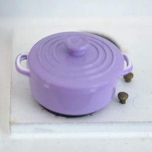 May include: A small purple enameled cast iron pot with white trim and two handles. The pot is sitting on a white surface.