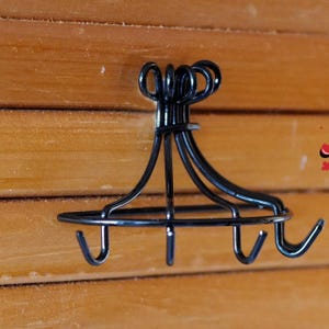 Op de afbeelding: Een zwarte metalen wandhaak met vier gebogen haken. De haak heeft een cirkelvormige basis en een decoratieve bovenkant met lusdetails. De haken zijn gelijkmatig verdeeld en steken naar buiten. De haak is op een houten oppervlak gemonteerd.