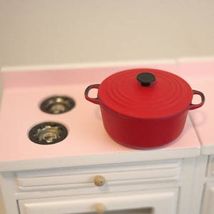 May include: A red enameled cast iron pot with a black knob on the lid sits on a pink countertop stovetop with two burner rings.