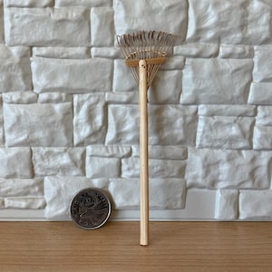 Peut inclure: Un petit râteau en bois avec un manche en bois brun et des dents en métal. Le râteau mesure environ 15 cm de long et est debout sur une surface en bois.