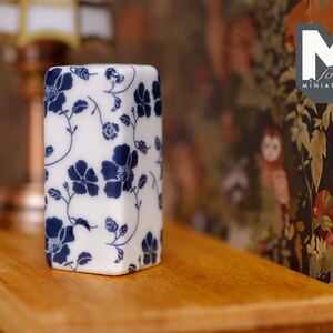 May include: A white ceramic decorative box with a blue floral pattern. The box is sitting on a wooden surface.